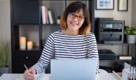 A professor sits at her desk in front of her computer with a notebook and smiles