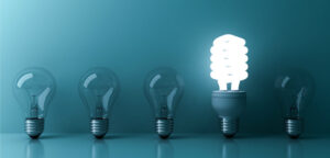 on a blue background, five lightbulbs in a row, four are traditional lightbulbs remain unlit, while the energy saving lightbulb is lit up.