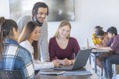 A small group of University students gather around an open laptop as they work together on a group project. They are each dresed casually and have smiles one their faces as they work away diligently.